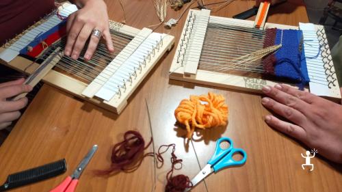 Mani al lavoro su due telai a cornice con filati colorati, spighe e fiori essiccati durante il laboratorio di tessitura a Bisaccia.
