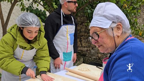 Una partecipante taglia la sfoglia su una spianatoia, mentre la cuoca locale e un altro partecipante osservano nel cortile a Castel Morrone.