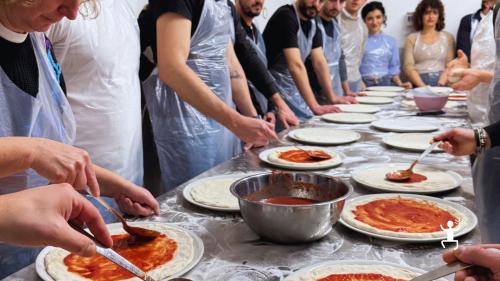 Partecipanti con grembiule distribuiscono la salsa di pomodoro sulle basi di pizza durante il laboratorio a Napoli.