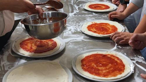 Mani dei partecipanti stendono il pomodoro sulle pizze disposte sul banco infarinato.