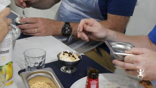 Mani di un partecipante che aggiunge gocce di cioccolato a una coppetta di gelato con granella durante il laboratorio a Napoli.