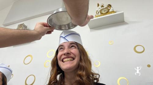 Partecipante sorridente con cappello da cuoco mentre una ciotola con crema viene capovolta sopra la sua testa durante il laboratorio di dolci a Napoli.