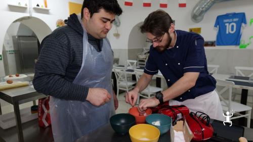 Chef affianca un partecipante nella preparazione delle uova in una ciotola, su un banco con ingredienti e fruste elettriche, durante il laboratorio a Napoli.